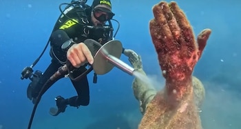 Buzo equipado con traje de neopreno y máscara que usa una herramienta para limpiar una estatua sumergida en el fondo del mar