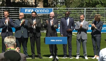 Siete personas vestidas de traje aplauden sobre un césped verde, con vallas azules que dicen "FIFA arena" de fondo. Hay un podio azul con "FIFA arena"