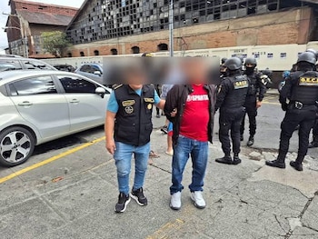 Dos hombres en primer plano, uno con chaleco, otro con camiseta roja. Múltiples agentes de seguridad con uniformes y cascos. Un coche blanco y edificios en el fondo