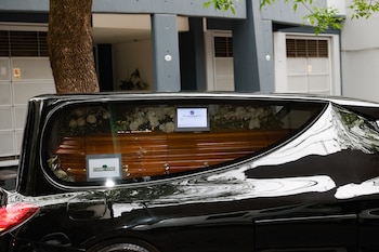 Un coche fúnebre negro lleva en su interior un ataúd de madera rodeado por flores blancas