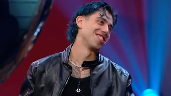 Luck Ra, un joven con cabello oscuro y chaqueta de cuero negra, sentado frente a un entrevistador de traje gris en un set de televisión con luces azules