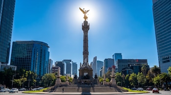 Monumento Ángel de la Independencia bajo un cielo azul claro en Ciudad de México. El sol brilla detrás de la estatua dorada. Edificios y tráfico vehicular rodean el área.