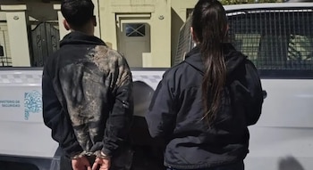 Un hombre de espalda con las manos esposadas permanece junto a una mujer con uniforme oscuro frente a un vehículo blanco