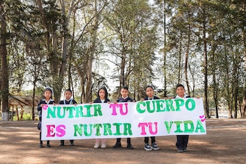 Seis niños uniformados sostienen un cartel horizontal blanco con letras verdes y rosadas que dice "Nutrir tu cuerpo es nutrir tu vida" frente a una arboleda