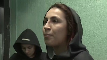 Primer plano de una mujer joven con cabello castaño oscuro y una sudadera con capucha negra, hablando frente a una pared verde texturizada