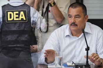Un hombre con bigote, camisa a rayas blanca, sentado en una mesa con un micrófono y un vaso de agua, con un chaleco de la DEA visible al fondo.