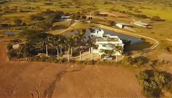 Vista aérea de una finca con una casa blanca, palmeras, un estanque y un coche blanco, rodeada de terreno árido y colinas