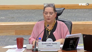Una mujer con suéter rosa habla ante un micrófono en una mesa con documentos, una computadora portátil y un cartel identificador