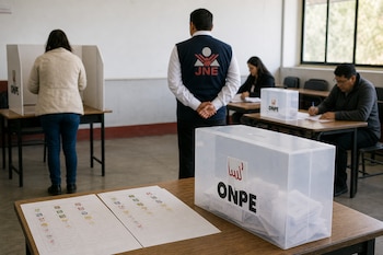 Una urna transparente con cédulas de sufragio sobre la mesa y personas en un aula de votación con un fiscalizador de espaldas.