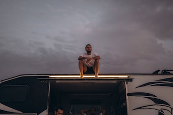 Hombre descalzo sentado sobre el techo de una autocaravana. Debajo, una luz horizontal sobre la puerta abierta y dos hombres parcialmente visibles. Cielo gris nublado