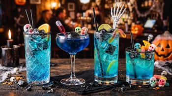 Cuatro vasos con bebidas azules, hielo negro, dulces de calavera y cítricos sobre una barra de madera con adornos de calaveras.