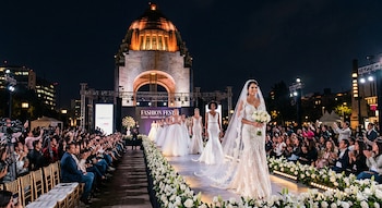 Desfile de moda nocturno en el Monumento a la Revolución, Ciudad de México. Modelos con vestidos de novia en pasarela florida, público sentado aplaudiendo