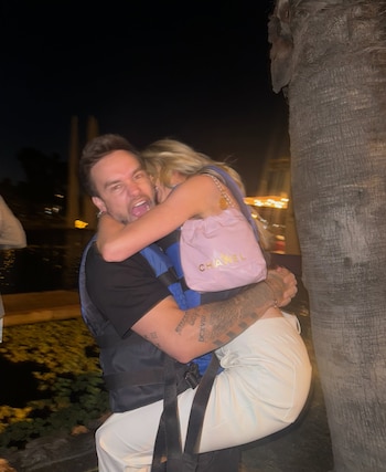 Un hombre con chaleco azul carga en brazos a una mujer con bolso rosado junto a una palmera de noche