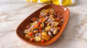 Plato ovalado de barro con patatas fritas, mejillones en conserva, tomate en cubos y cebolla morada sobre una superficie de mármol.