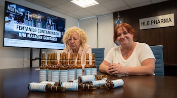 Dos mujeres sentadas frente a una mesa con varias ampollas de fentanilo. Detrás, una pantalla exhibe un titular sobre fentanilo contaminado y un cartel de HLB Pharma.