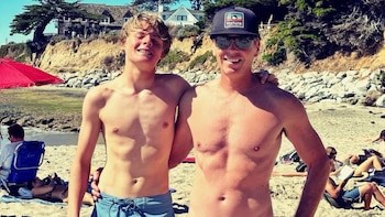Dos hombres sonrientes y sin camiseta posan en una playa de arena. Al fondo hay un acantilado con vegetación, casas, sombrillas y personas