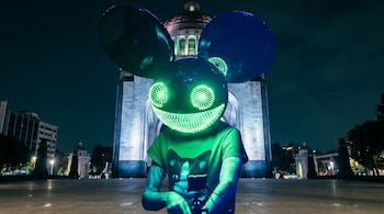 Una persona con una máscara negra de grandes orejas y ojos y boca de luz verde, vistiendo una camiseta verde, de pie frente al iluminado Monumento a la Revolución de noche.