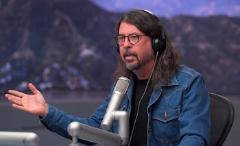 Dave Grohl, con cabello largo, barba, gafas y auriculares, usa una chaqueta vaquera mientras habla a un micrófono plateado sobre una mesa. Fondo montañoso borroso