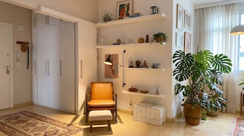Sala de estar con armario blanco, sillón de cuero y otomana, estantes flotantes con plantas y objetos decorativos, planta Monstera, cortinas y alfombras.
