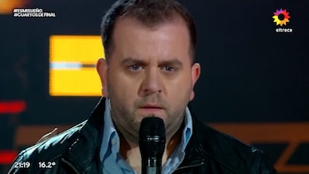 Hombre con chaqueta negra y camisa azul, sosteniendo un micrófono negro. Fondo oscuro con luces amarillas. Logos de canal eltrece y programa Es Mi Sueño