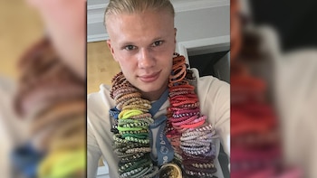 Un hombre joven con pelo rubio posa de frente con múltiples gomitas de pelo de colores alrededor del cuello, vestido con una prenda blanca