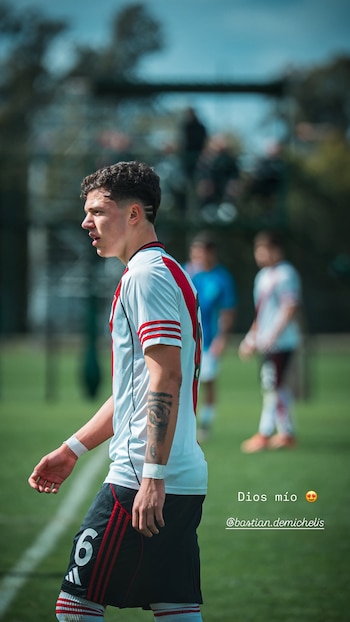 Un joven futbolista con camiseta blanca de franja roja y pantalón negro camina de perfil sobre el césped de una cancha