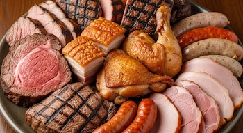 Primer plano de un gran plato rebosante de diferentes carnes cocinadas, incluyendo rosbif, trozos de panceta crujiente, pollo, salchichas y jamón.