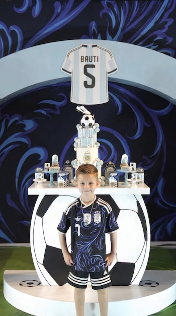 Niño con camiseta de la selección argentina posa frente a una decoración de cumpleaños con la camiseta número cinco, una torta y balones de fútbol.