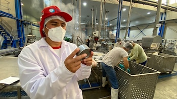 Un hombre con bata, mascarilla y gorro examina una lata de metal en una fábrica de alimentos con operarios y maquinaria industrial