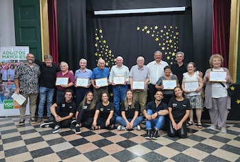 Un grupo de adultos y personas mayores posa en un escenario con diplomas enmarcados, un telón negro con estrellas y un cartel institucional