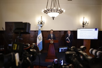 Hombre con traje y gafas habla en un podio. Bandera de Guatemala a la izquierda, pantalla digital con documentos a la derecha. Fondo con paneles de madera