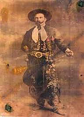 Fotografía antigua sepia de un hombre con sombrero, pañuelo, chaqueta y pantalón bordados, y cinturón adornado, de pie en un estudio, barbudo