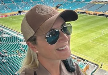 Mujer con gorra marrón, gafas de sol y uniforme beige. Al fondo, un estadio de fútbol con campo de juego verde y gradas con asientos