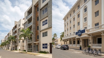 Vista urbana de República Dominicana con edificios modernos de alquiler temporal a la izquierda y un hotel tradicional de varios pisos a la derecha, con palmeras, autos y bicicletas.