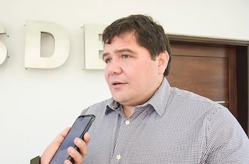 Un hombre con camisa a cuadros sostiene un teléfono móvil negro. Se observan letras "S", "D", "E" en una pared blanca y una puerta de madera