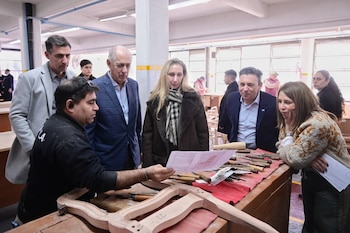 Un hombre muestra un plano de diseño de una pieza de madera a un grupo de cinco personas en un taller con herramientas de carpintería