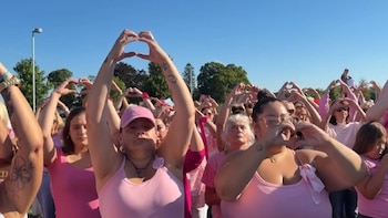 Varias personas con ropa rosa forman figuras de corazones con sus manos en un espacio exterior bajo un cielo despejado