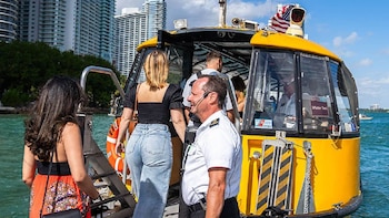 Vista lateral de un Water Taxi amarillo en Miami Beach con pasajeros abordando. Un tripulante en uniforme blanco supervisa, con edificios altos al fondo y cielo azul