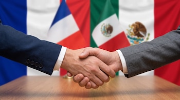 Dos manos en traje formal se estrechan sobre una mesa de madera, con las banderas de Francia y México al fondo.