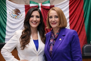 Kenia López Rabadán con blazer blanco y Laura Itzel Castillo con blazer morado, sonriendo y posando frente a la bandera de México.