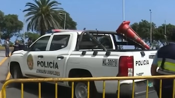 Camioneta blanca de la Policía con luces de emergencia y texto 'Policía Comisaría Miraflores', un cono rojo en la parte trasera, personas y palmeras