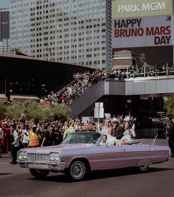 Coche convertible rosa con gente en desfile de Las Vegas. Una multitud. Cartel del Park MGM: 'Happy Bruno Mars Day' y foto de Bruno Mars