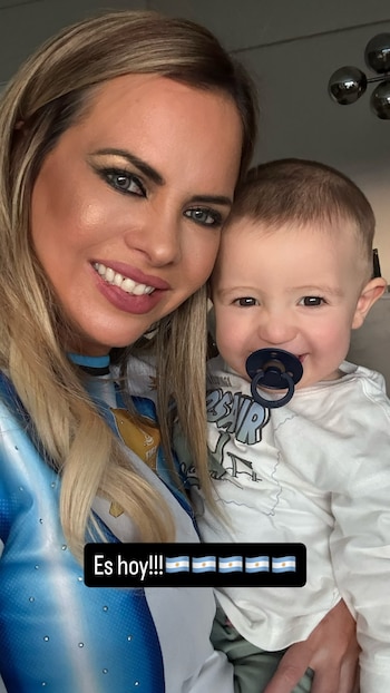 Alejandra Maglietti sonríe y sostiene a un niño con chupete. Ambos visten camisetas celestes y blancas. Hay un cartel negro que dice "Es hoy!!!" y varias banderas de Argentina