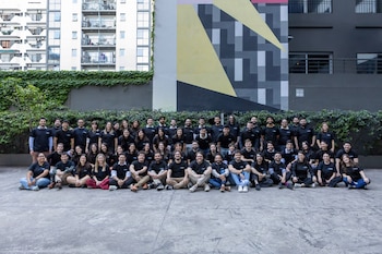 Un grupo de aproximadamente sesenta personas en camisetas negras posa al aire libre frente a un muro con hiedra, un mural geométrico colorido y edificios modernos