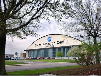 Un gran hangar blanco con el logotipo de la NASA y el texto Glenn Research Center, rodeado de árboles, césped y automóviles estacionados