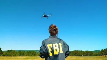 Persona de espalda con chaqueta negra con letras amarillas 'FBI' mira un dron volando sobre un campo verde bajo cielo azul