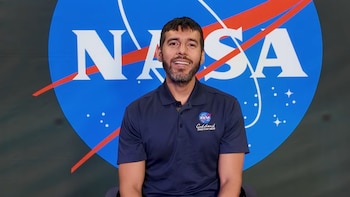 Un hombre sonriente con barba y camiseta polo azul marino con el logo de NASA Goddard. De fondo, un gran logo azul de la NASA