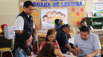 Un grupo de personas sentadas en un aula revisa documentos sobre las mesas junto a una cabina de votación y una caja electoral.