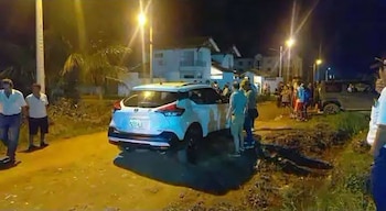 Un automóvil blanco permanece estacionado en una calle de tierra iluminada por farolas nocturnas junto a varias personas reunidas