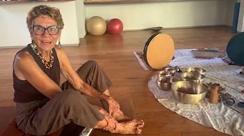 Mujer de cabello rizado con gafas y blusa oscura, sentada en un tapete de yoga, con cuencos tibetanos, tambor de marco y pelotas de ejercicio.